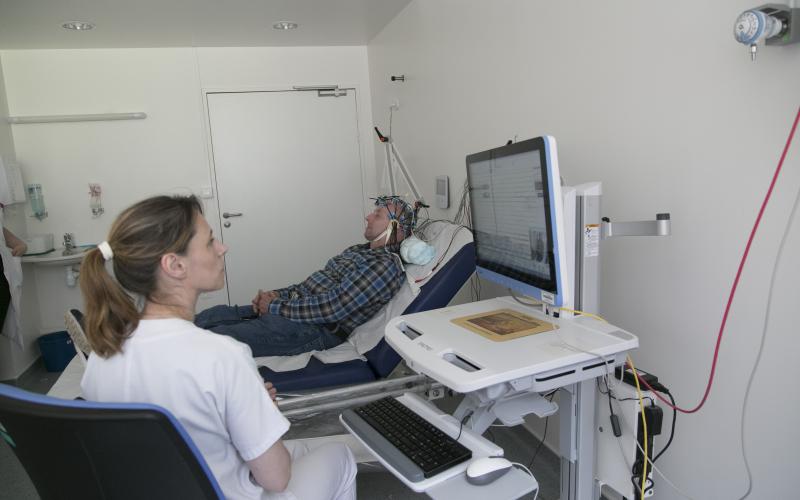 un homme allongé avec une machine sur la tête et une infirmière qui  regarde des résultats sur son ordinateur 
